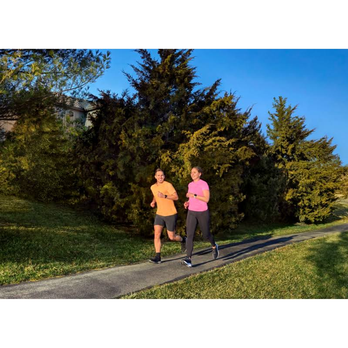 Dwóch biegaczy na ścieżce z zielenią i drzewami. Jeden ma pomarańczowy, drugi różowy. Błękitne niebo w tle.