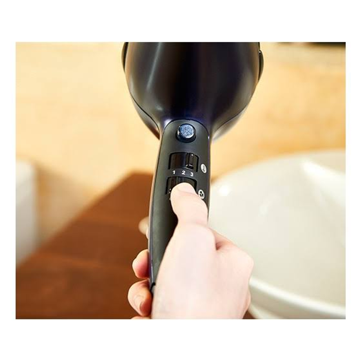 Dłoń trzyma czarną suszarkę, reguluje prędkość. Beżowa ściana i biała umywalka w tle.