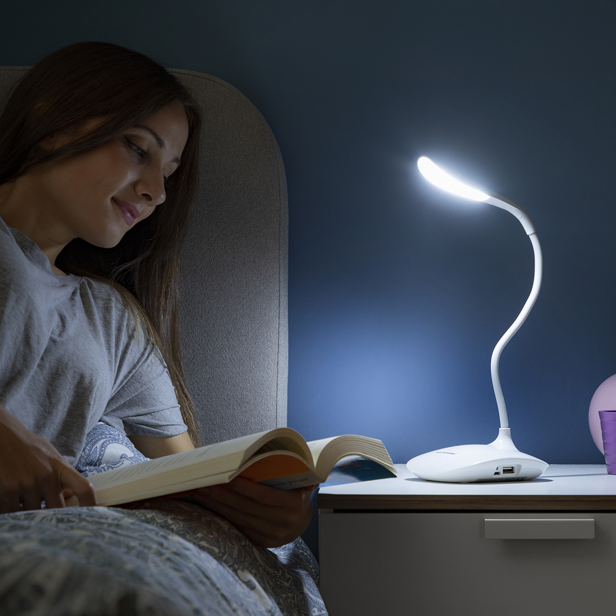 Donna che legge un libro sotto una lampada da scrivania bianca. La lampada è su un comodino.