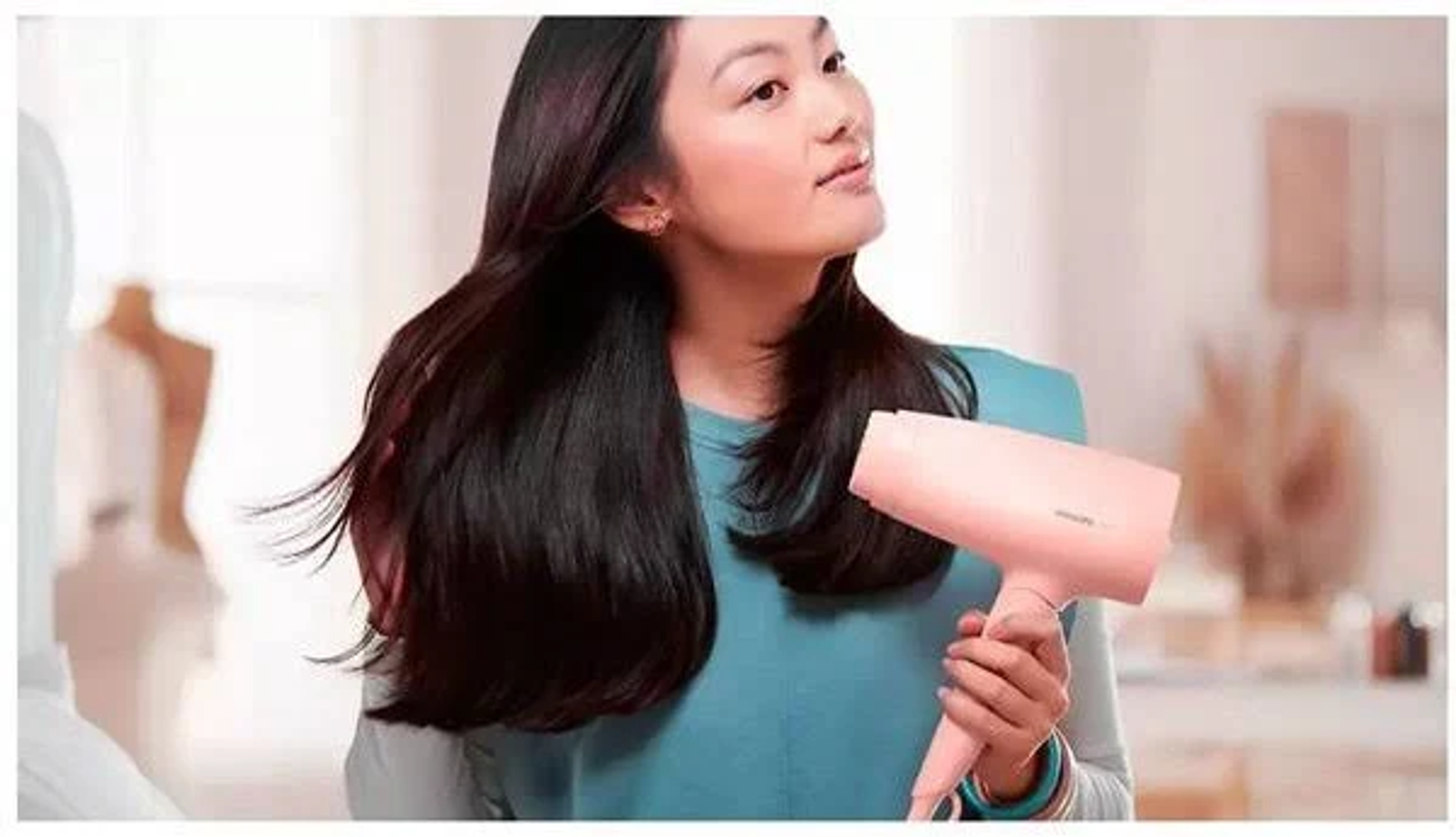 Donna con lunghi capelli scuri che usa un asciugacapelli rosa. Indossa un top verde acqua. Sfondo sfocato.