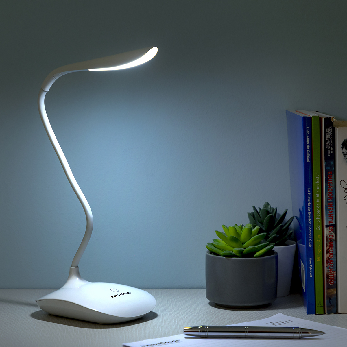 Lampada da scrivania bianca con collo curvo, che brilla su una scrivania con libri e piante.