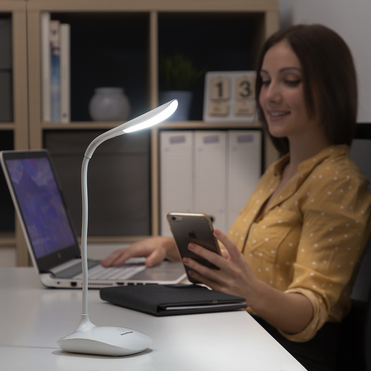 Lampada da scrivania bianca su una scrivania. Donna che usa laptop e telefono. Scrivania bianca, sfondo scuro.