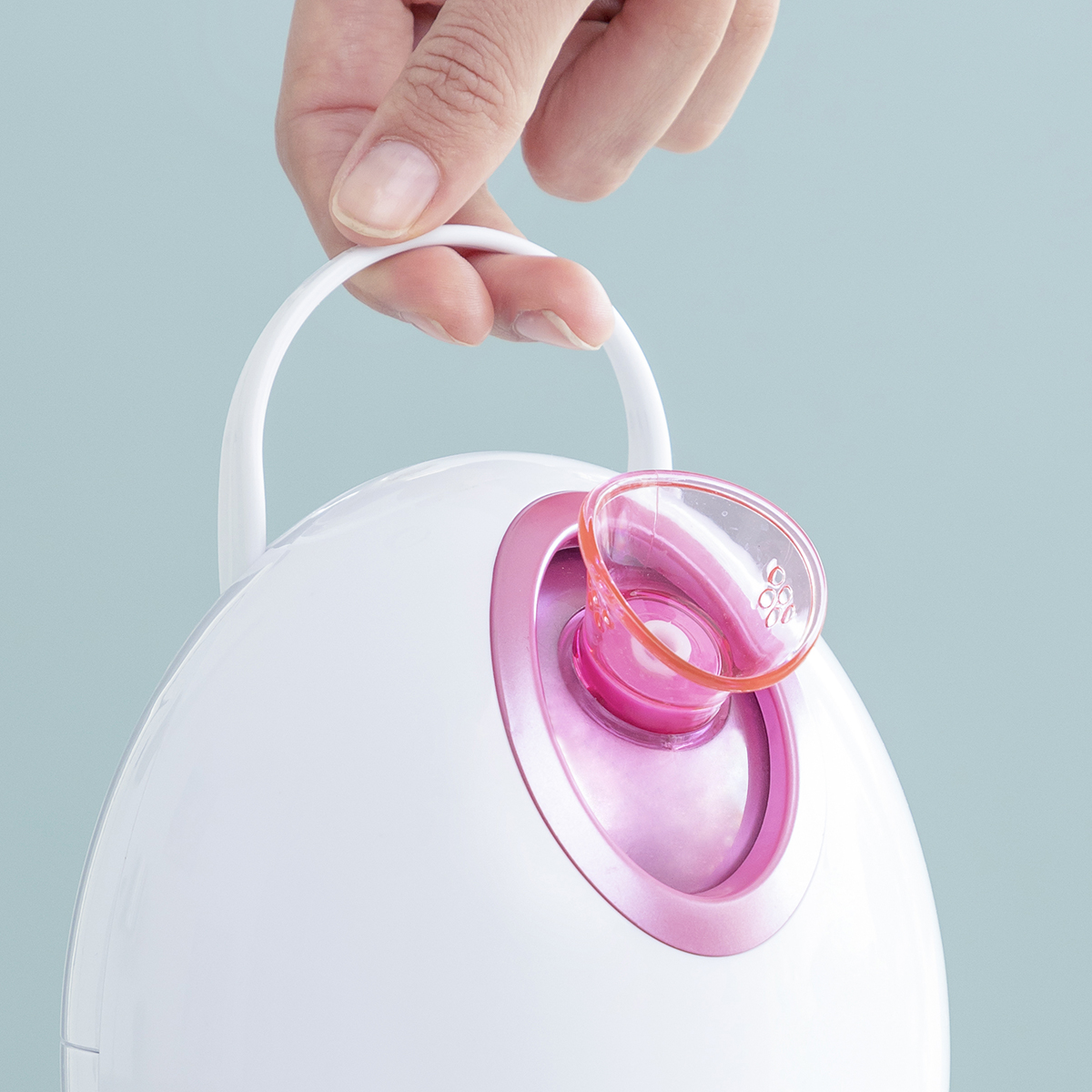 Un vaporizzatore per il viso bianco. Una mano tiene la maniglia. Il vaporizzatore ha un interno rosa e un beccuccio trasparente.