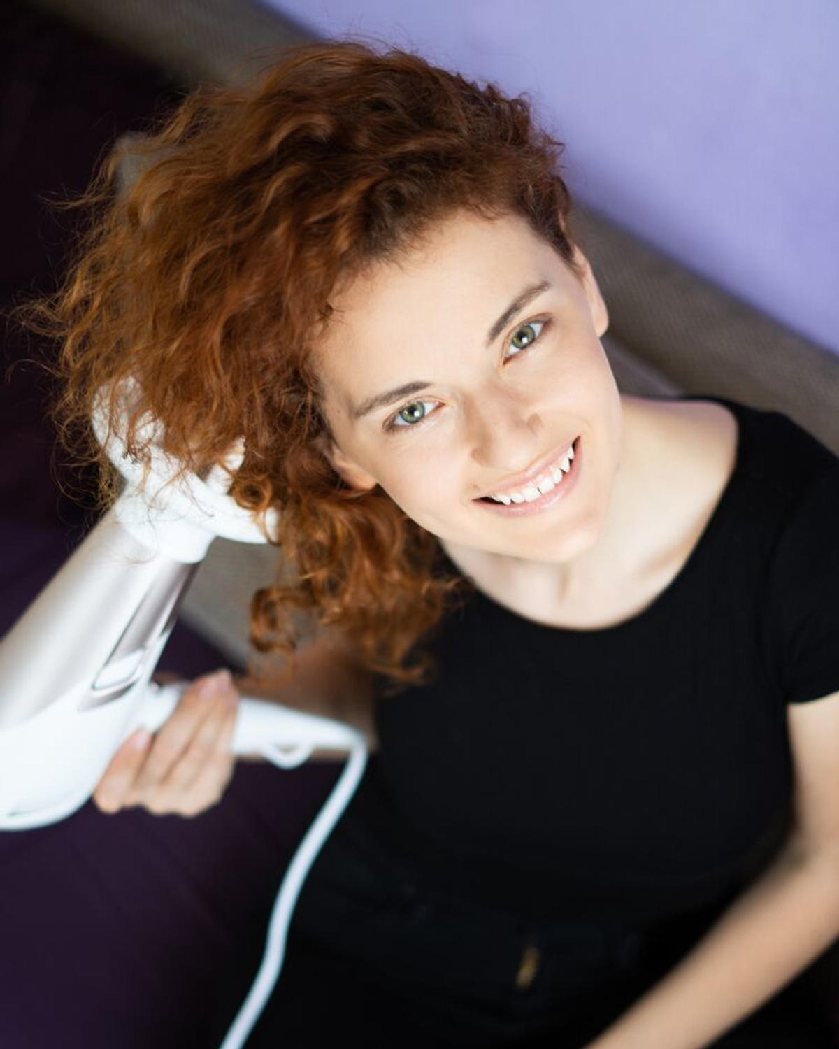 Uśmiechnięta kobieta z rudymi kręconymi włosami suszy włosy białą suszarką, patrząc w górę. Fioletowe tło.