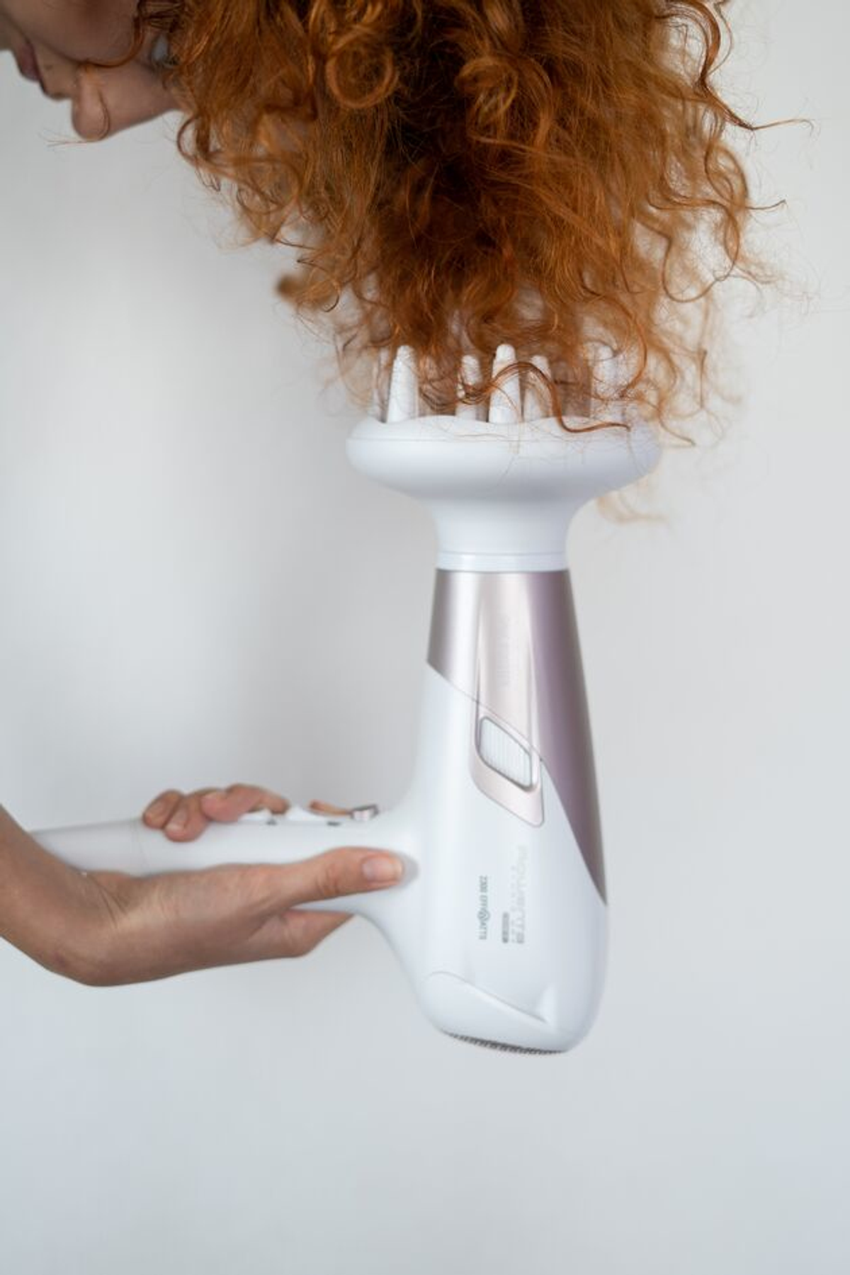 Osoba trzyma białą i fioletową suszarkę do włosów, susząca czerwone kręcone włosy.
