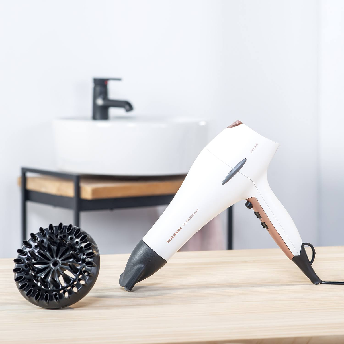 Secador de pelo blanco y oro rosa con difusor desmontado sobre una superficie de madera, cerca de un lavabo.
