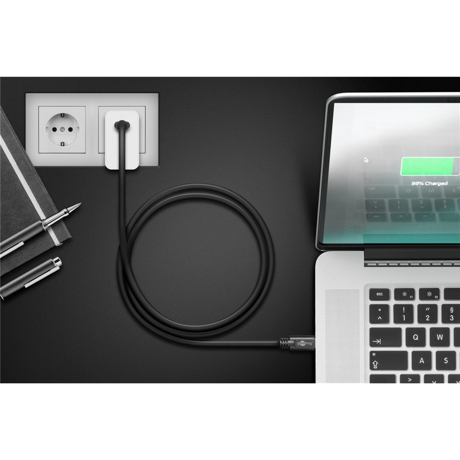 Laptop in carica tramite un cavo nero da una presa a muro, con penna e quaderno nelle vicinanze.