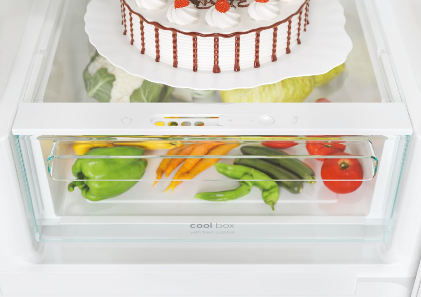 Frigorifero con torta, verdure. Torta sullo scaffale superiore, verdure nel cassetto sottostante, sfondo bianco.