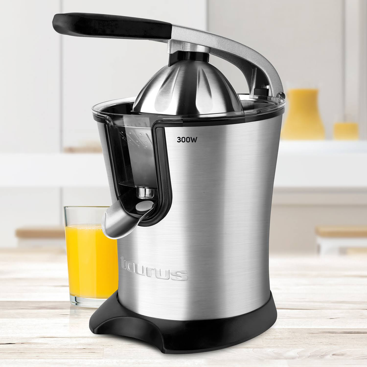 Una exprimidora eléctrica plateada y negra con un vaso de zumo de naranja y fondo blanco.