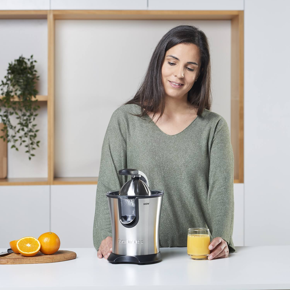 Mujer exprimiendo naranjas con un exprimidor eléctrico, sosteniendo un vaso.