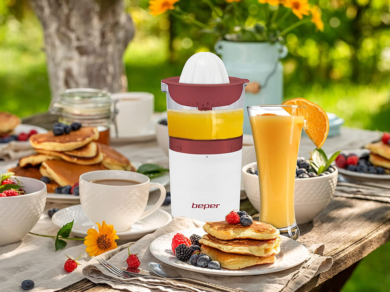 Un tavolo apparecchiato per la colazione, con spremiagrumi, succo, pancake, frutti di bosco, caffè e un fiore.