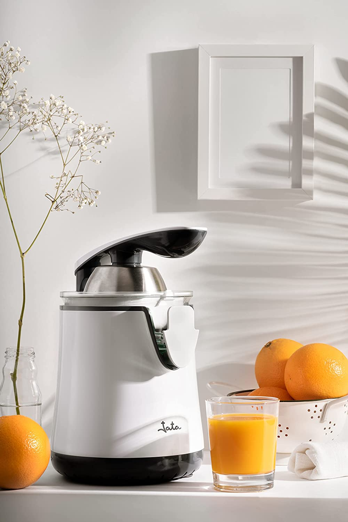 Un exprimidor blanco y negro sobre un estante blanco con naranjas, un vaso de zumo y un cuadro enmarcado.