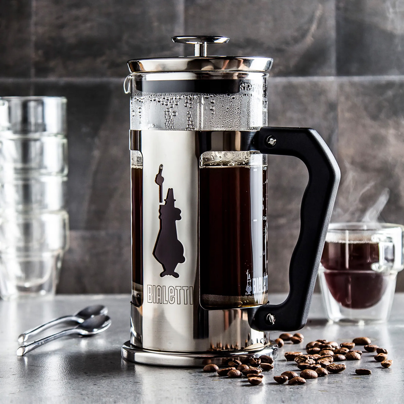 Eine French-Press-Kaffeemaschine mit Kaffee, Kaffeebohnen und einer Tasse Kaffee. Der Hintergrund ist grau.