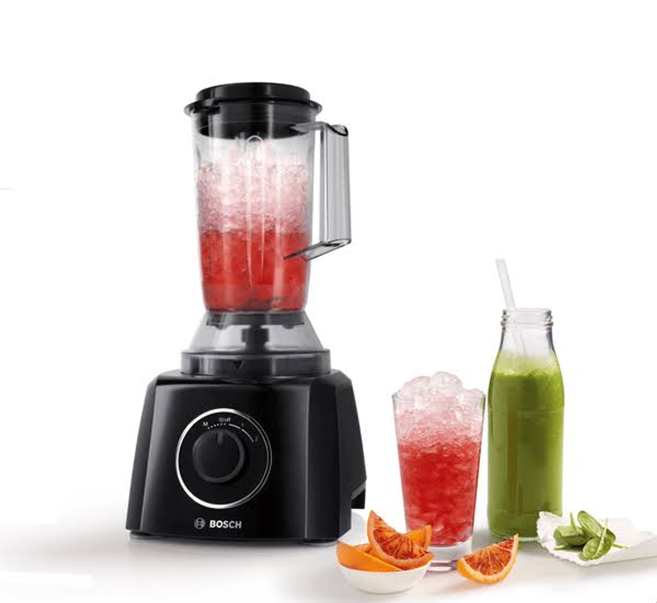 Czarny blender z pojemnikiem wypełnionym czerwoną cieczą i lodem, obok szklanki i butelki zielonego soku.