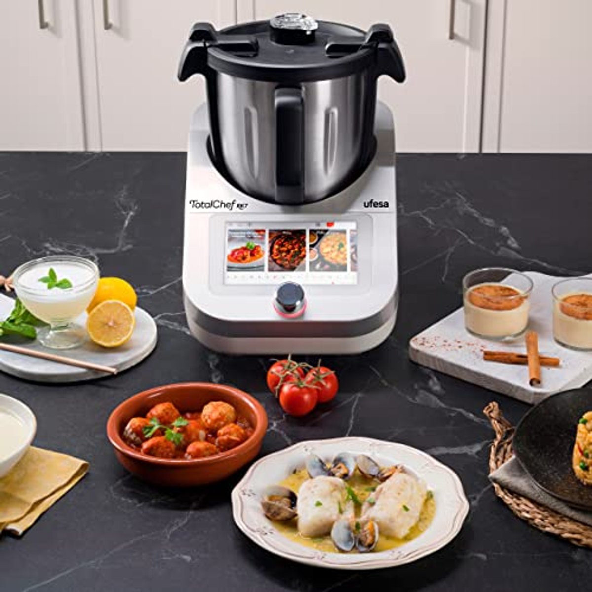 Eine Küchenmaschine, ein Tisch mit Essen darum. Ein weißes, graues und schwarzes Gerät mit Bildschirm.
