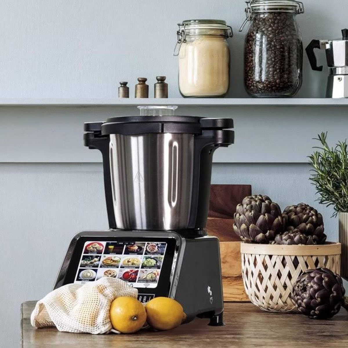 Un electrodoméstico de cocina negro con pantalla táctil, limones y alcachofas sobre una mesa frente a una estantería.