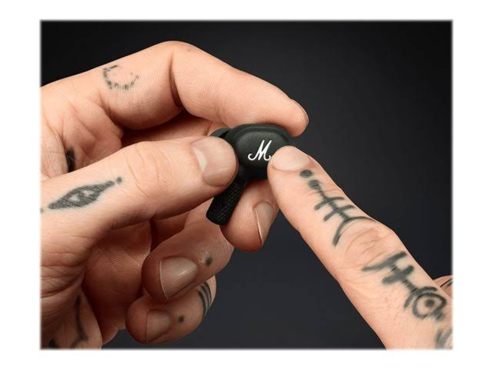 Hände einer Person, die einen schwarzen Ohrhörer mit weißem 'M'-Logo halten, mit Tattoos auf den Fingern und Armen.