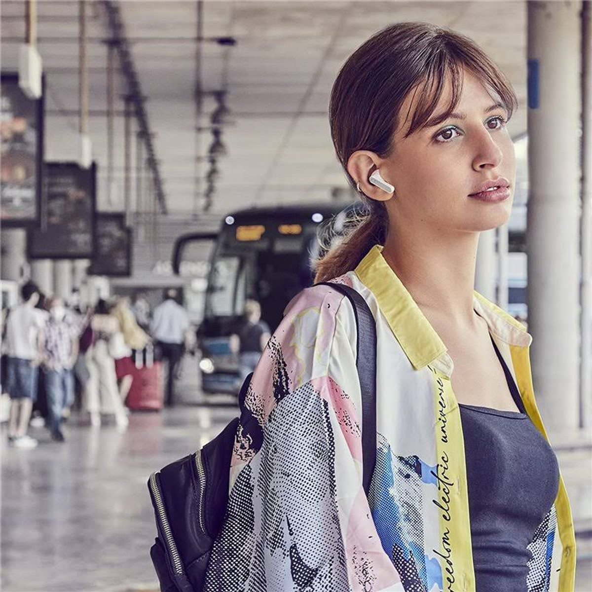 Una mujer con auriculares, de pie en una estación con una mochila. Mira hacia otro lado.