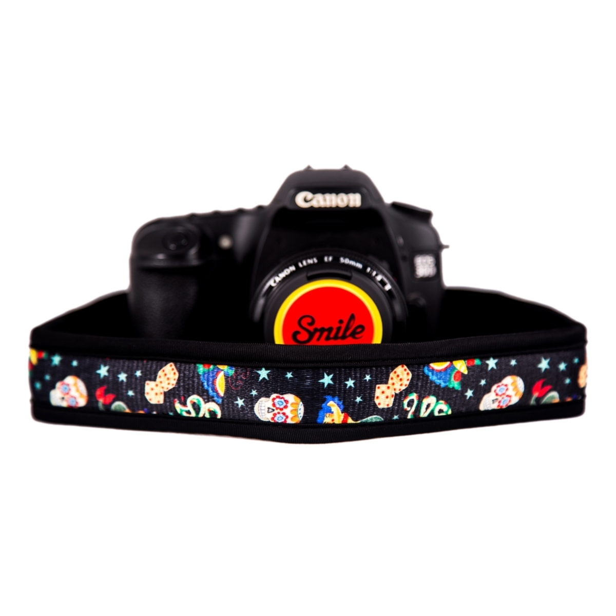 Fotocamera Canon con tappo obiettivo sorriso. Cinturino nero con motivi colorati su bianco.