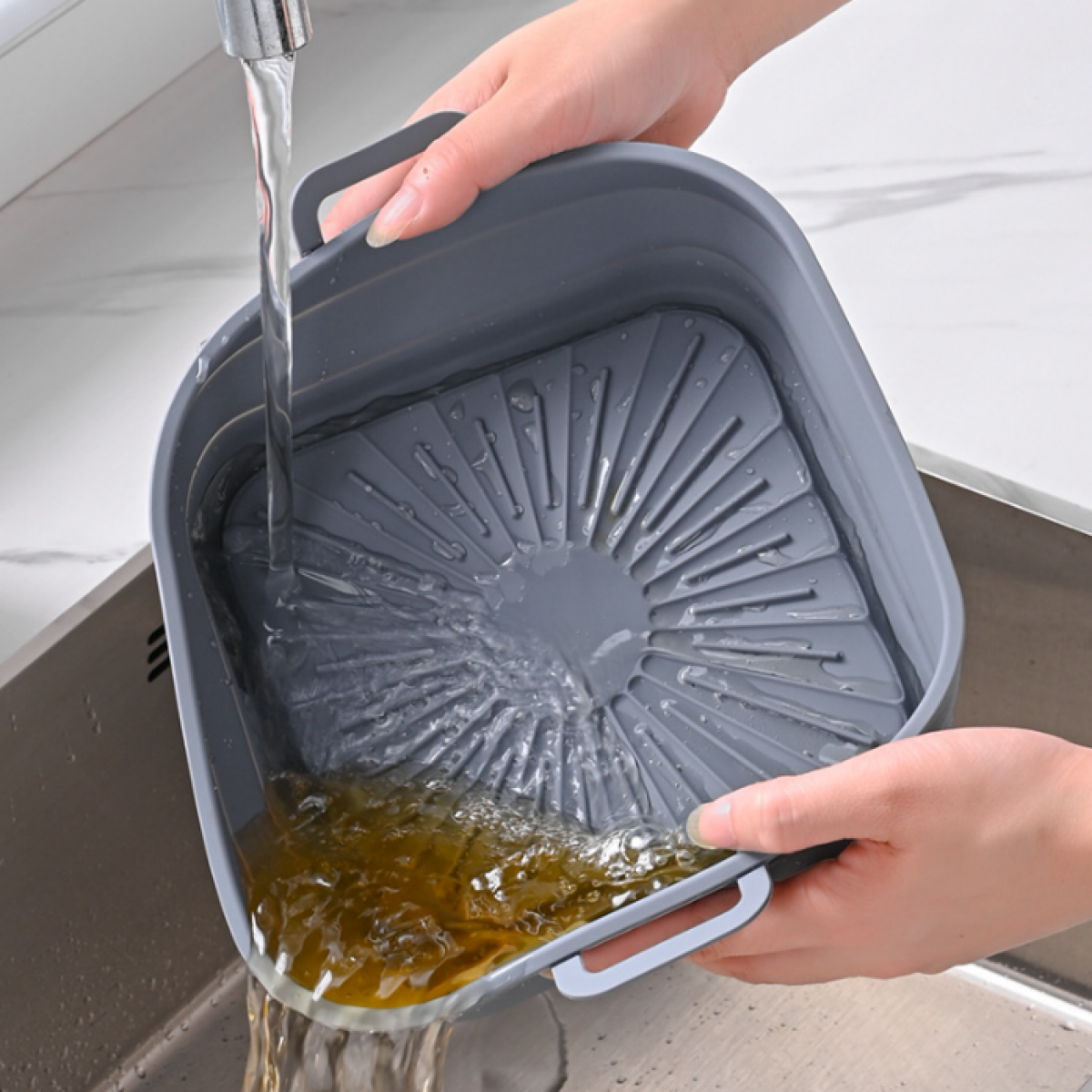 Handen wassen een grijze, vierkante siliconen zeef onder stromend water, met eten erin.