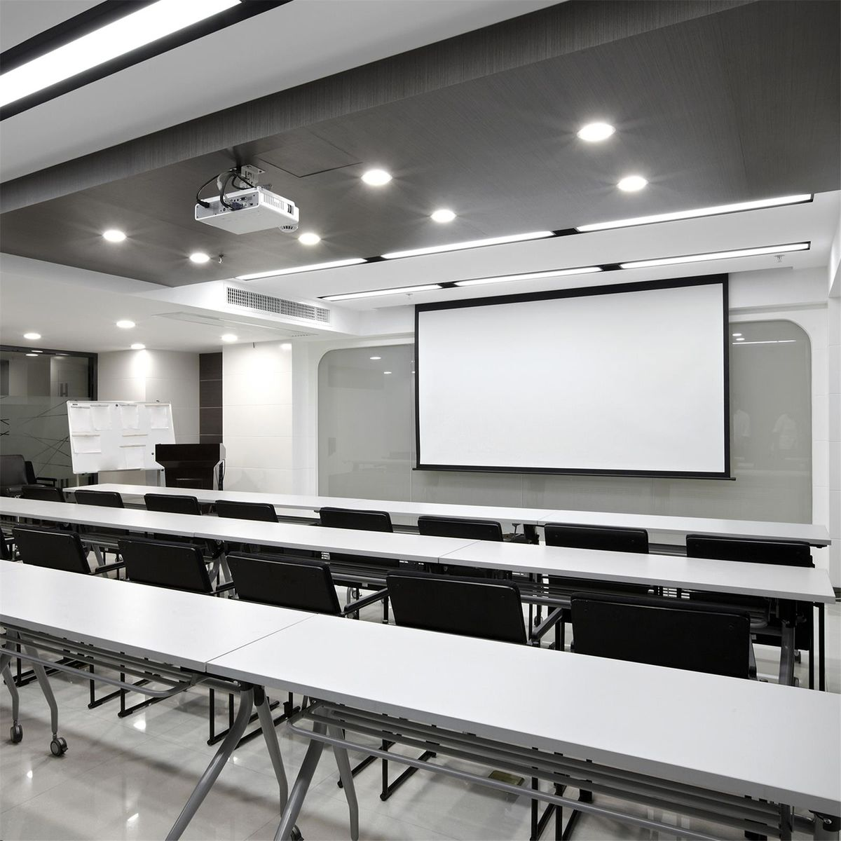 Een vergaderzaal met witte tafels, zwarte stoelen, een projector en een wit scherm.