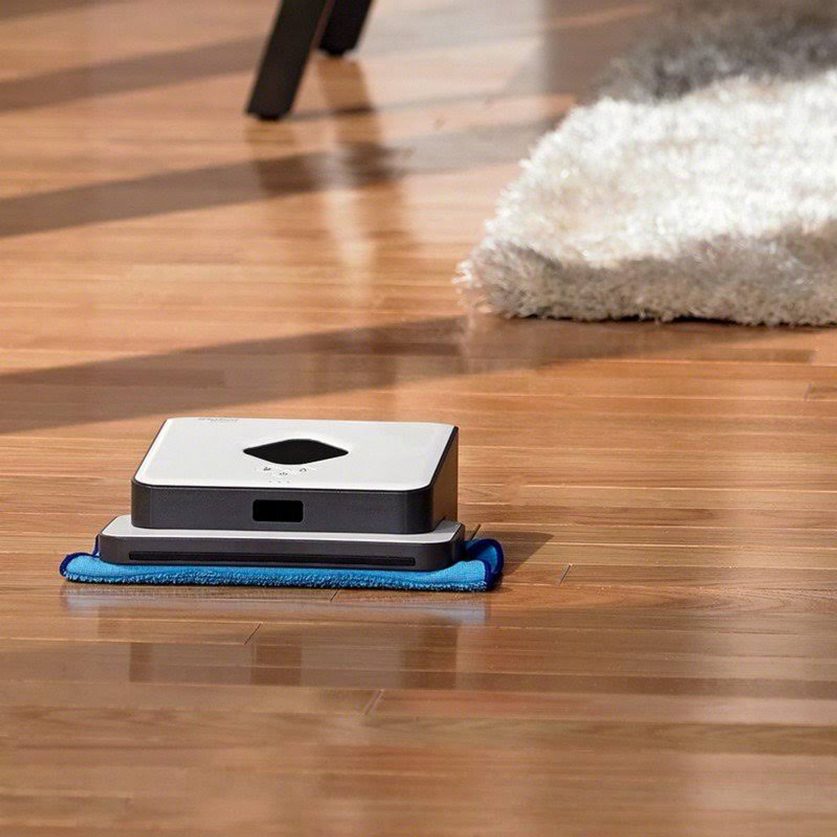 Roboter-Staubsauger auf Holzboden, blaues Pad und ein weißer Teppich im Hintergrund.