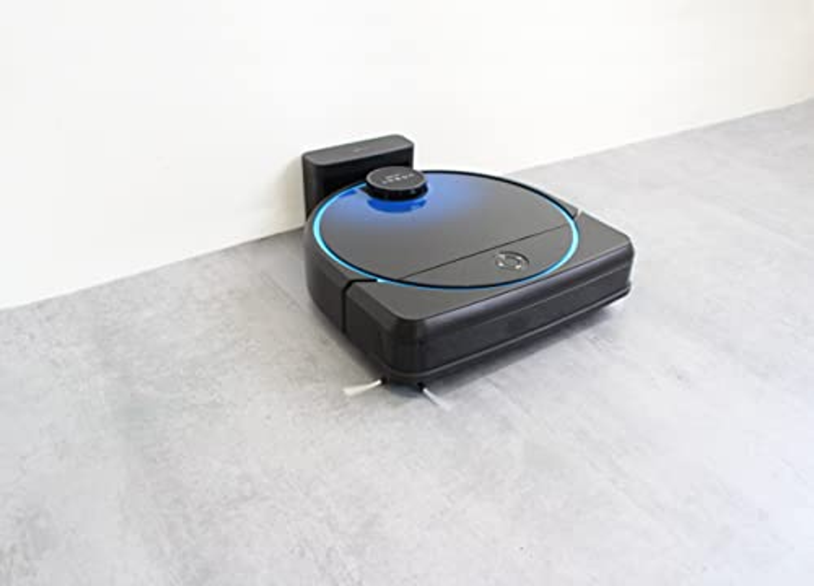Una aspiradora robot negra y azul sobre un suelo gris, cerca de una pared blanca.