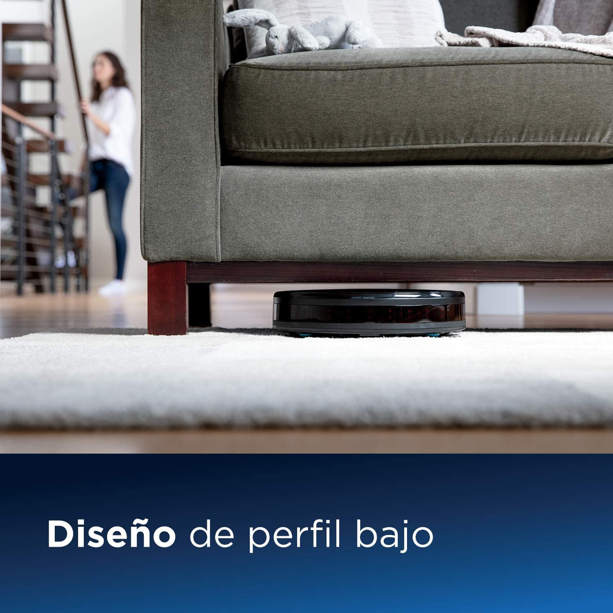 Robot aspirador debajo de un sofá. Mujer al fondo. Texto: Diseño de perfil bajo.