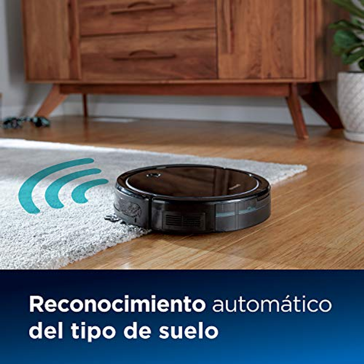 Una aspiradora robot negra sobre una alfombra, con un armario de madera en el fondo.