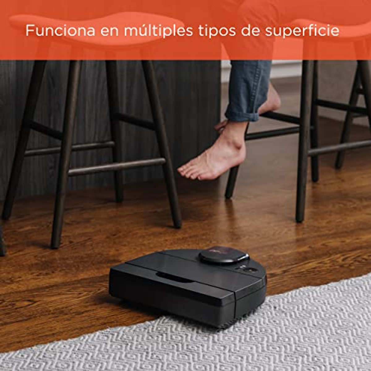 Ein schwarzer Roboter-Staubsauger auf einem Holzboden. Füße und Beine einer Person sind im Hintergrund.