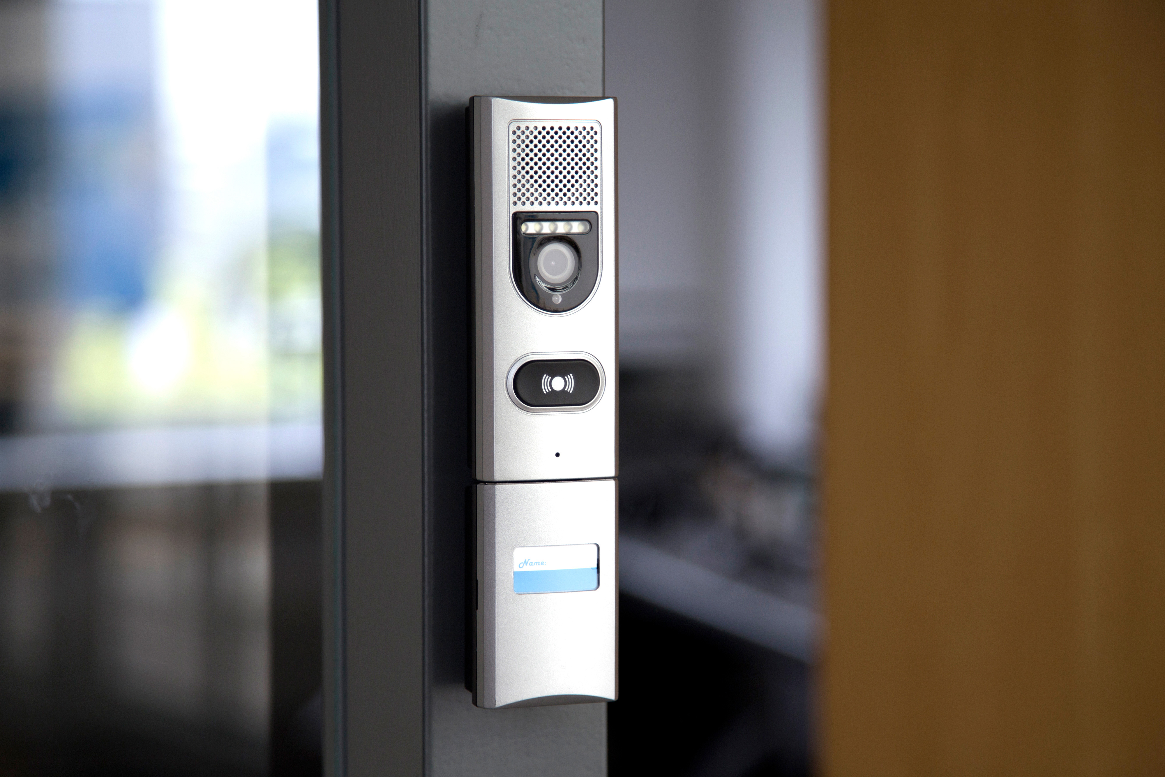 Silberne intelligente Türsprechanlage an einer Wand, Kamera und Knopf sichtbar, mit einer Glas-Tür-Reflexion.