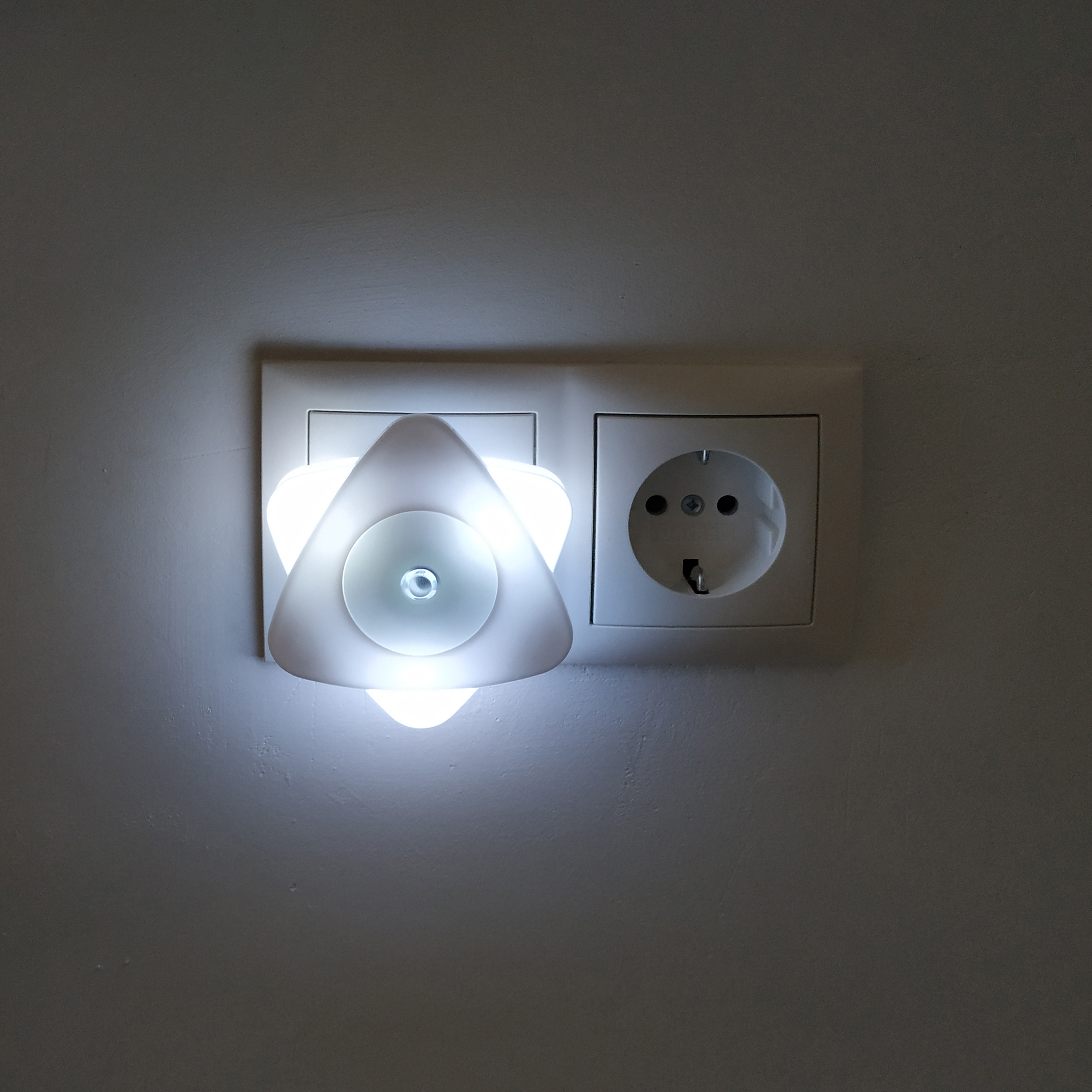 Biała, podświetlana trójkątna lampka nocna wpięta do gniazdka ściennego.