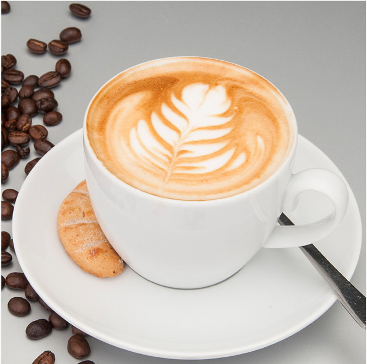 Latte in einer weißen Tasse mit Blattkunst, Untertasse, Keks und Kaffeebohnen auf grauem Untergrund.