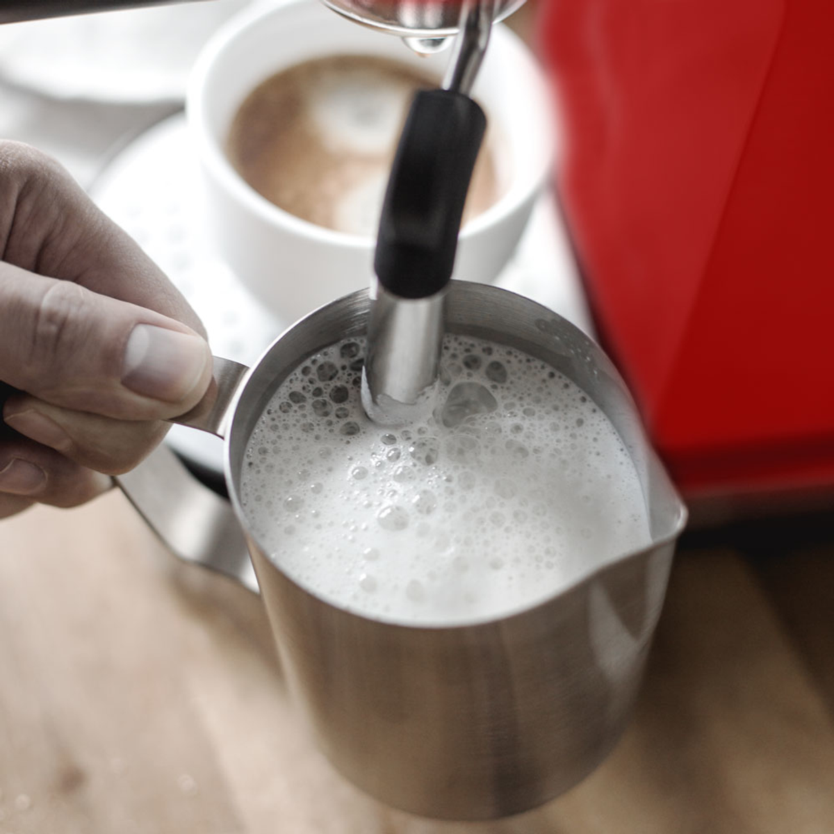 Ręka spieniająca mleko w metalowym dzbanku. Filiżanka kawy i czerwony ekspres do kawy w tle.