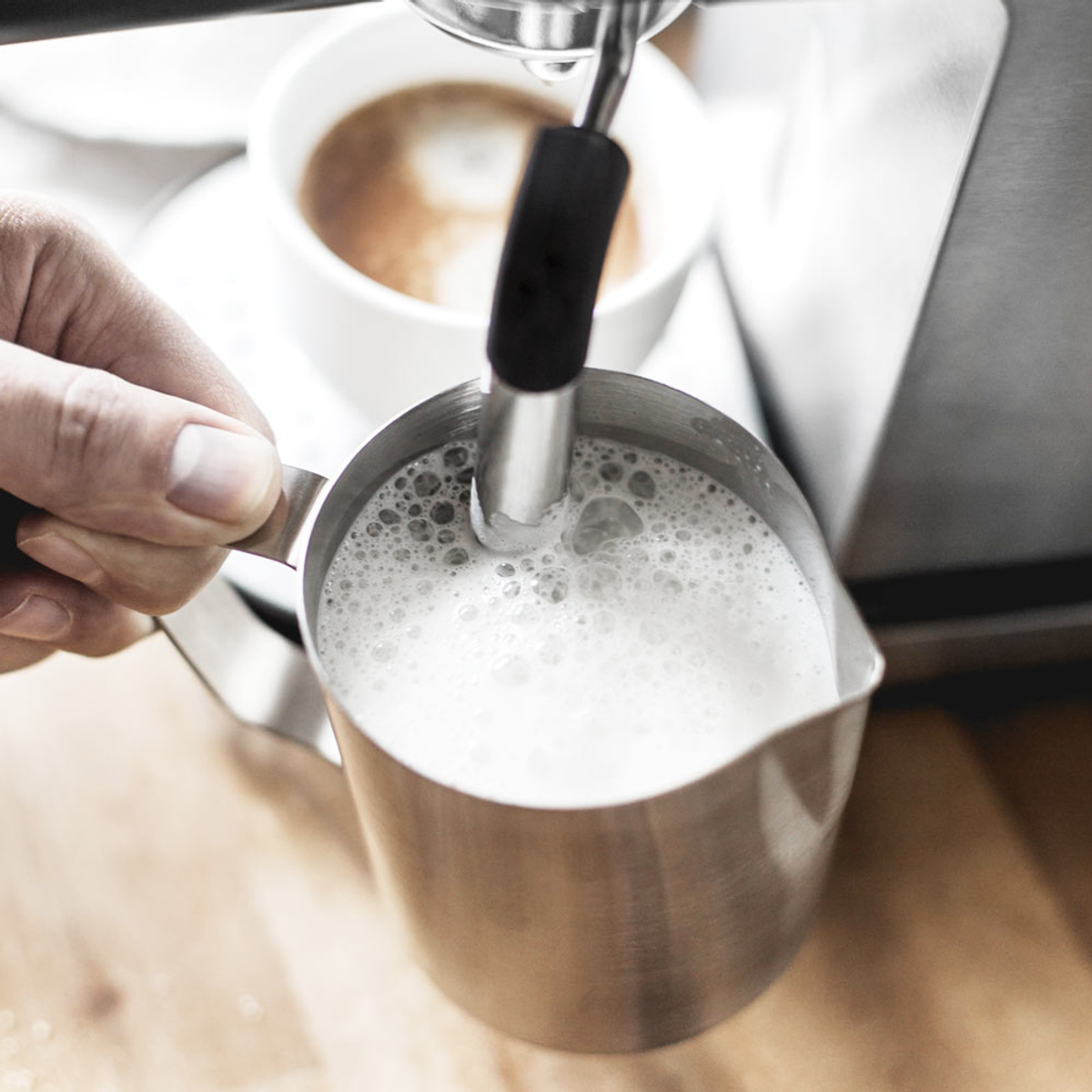 Osoba robiąca latte art, wlewa mleko do srebrnego dzbanka. Filiżanka kawy w tle.