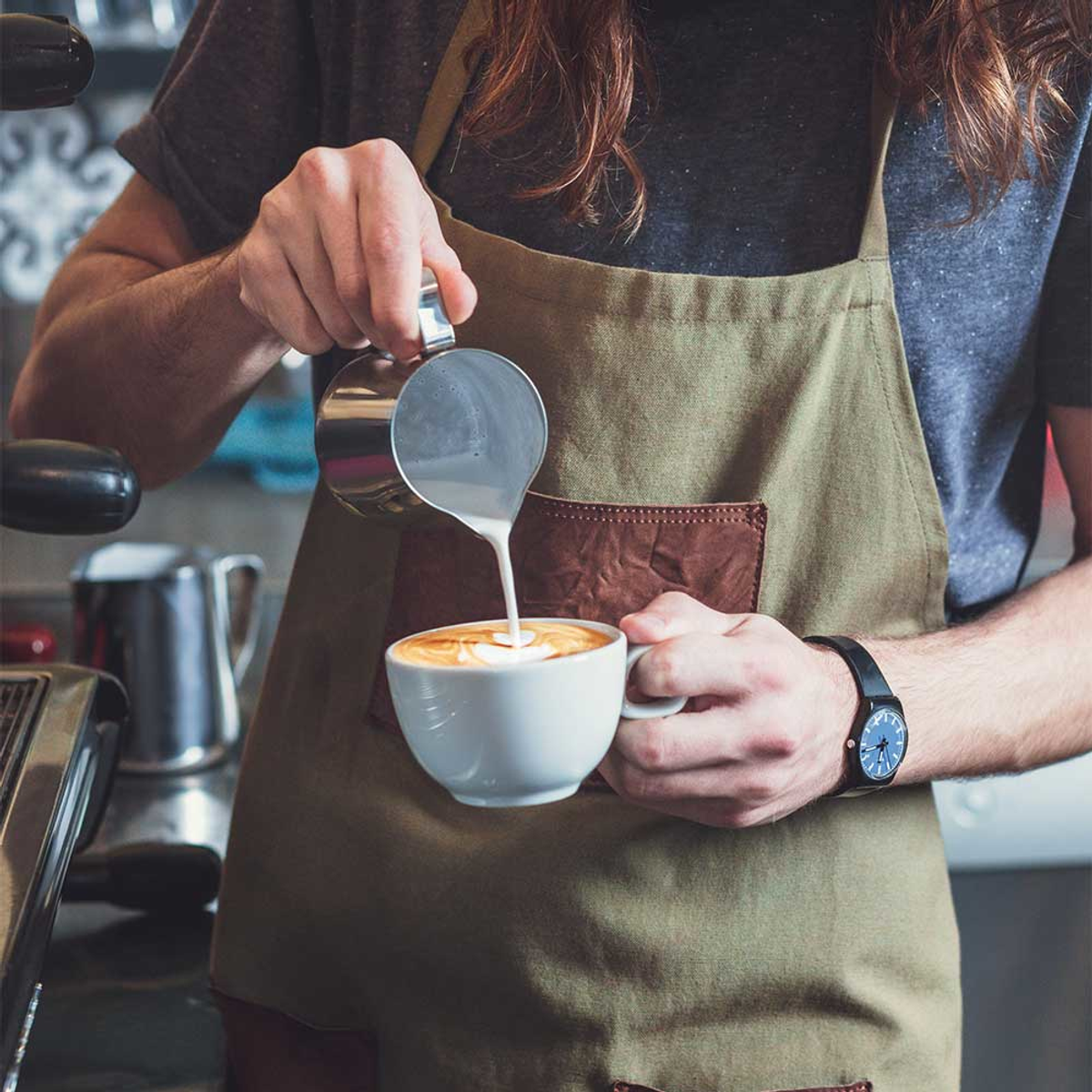 Barista nalewa mleko z metalowego dzbanka do białej filiżanki kawy z latte art.