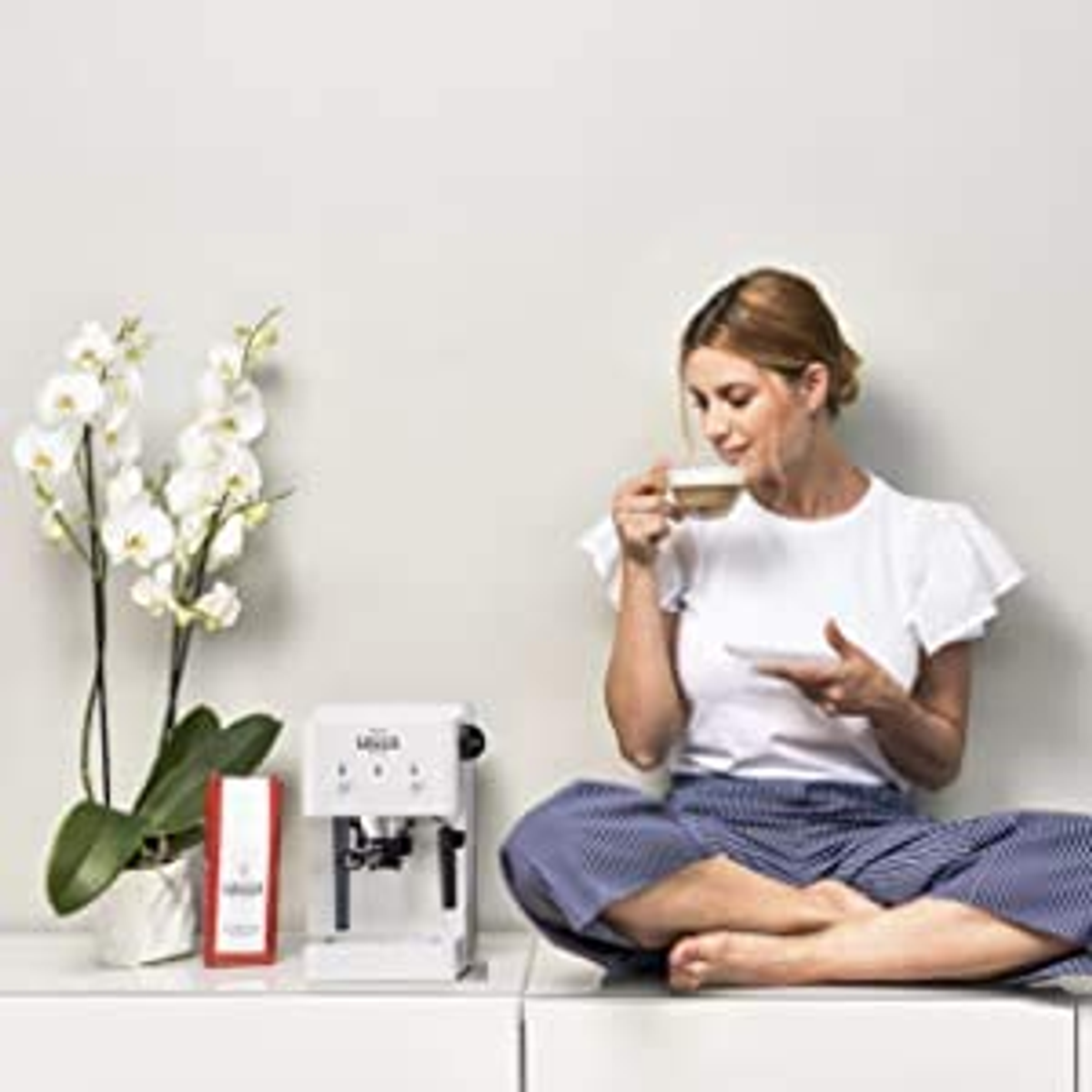 Una mujer sentada, bebiendo café, cerca de una máquina de espresso blanca y orquídeas en un estante.