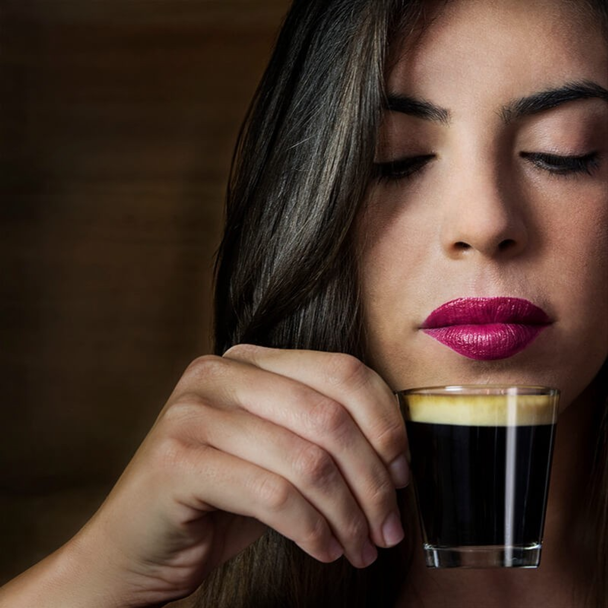 Frau mit rotem Lippenstift hält ein Glas Kaffee, Augen geschlossen.
