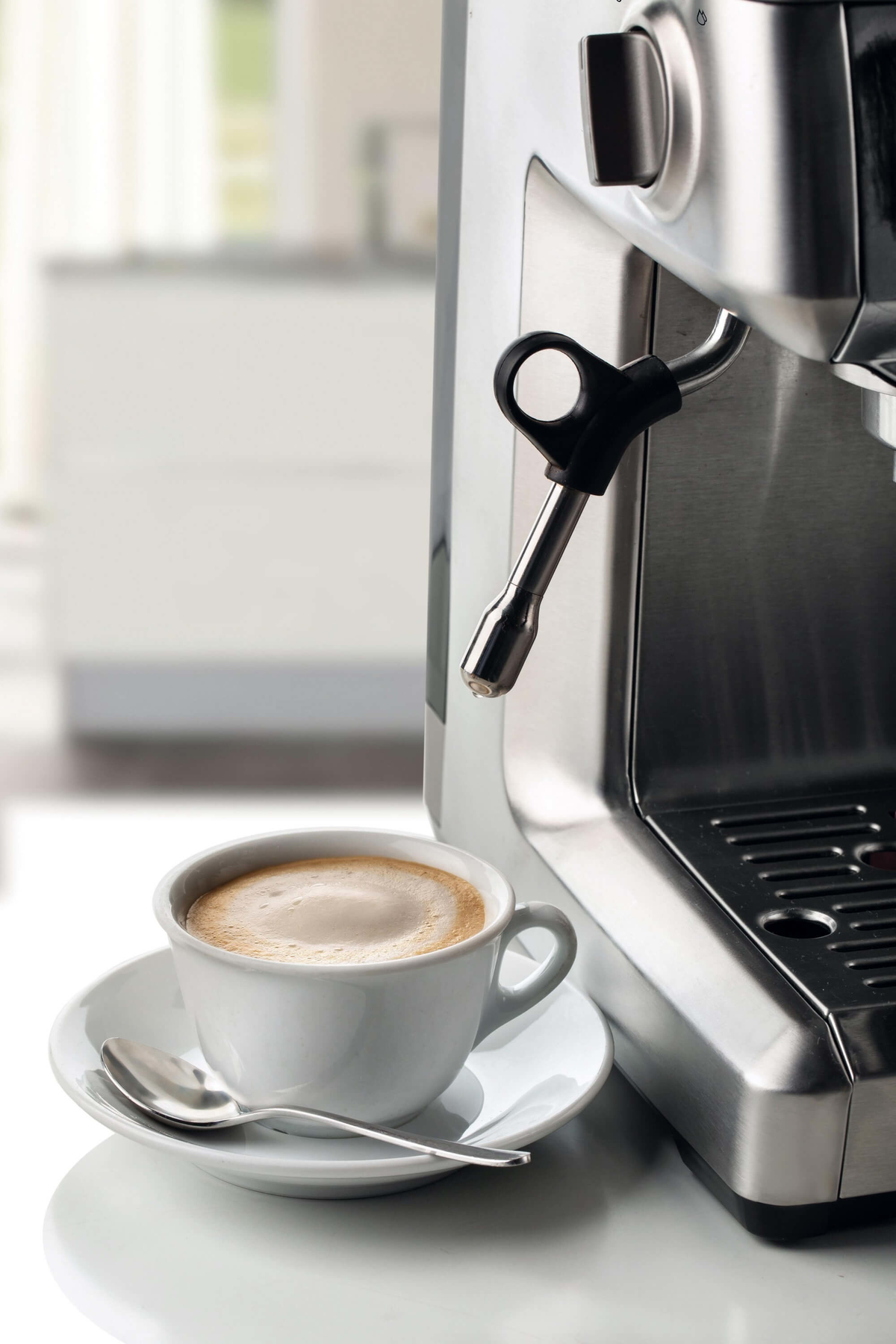 Máquina de espresso y una taza blanca con café en un platillo. Una cuchara plateada está en el platillo.