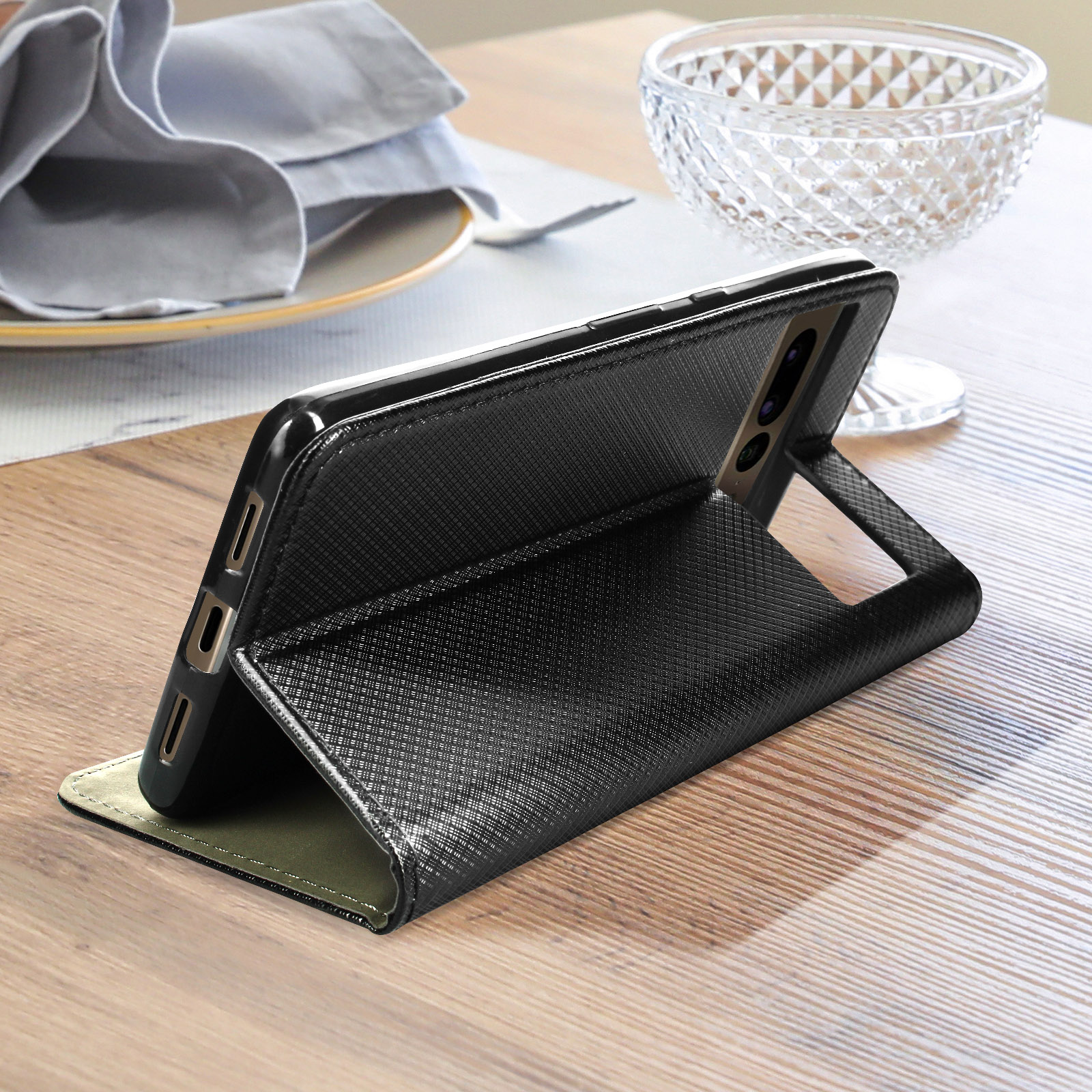 Custodia per telefono nera con design strutturato, tiene un telefono su un tavolo di legno, bicchiere e stoffa sullo sfondo.