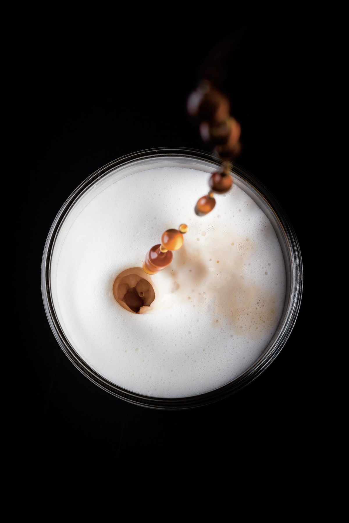 Kawa wlewana do szklanki z góry, na czarnej powierzchni. Biała piana.