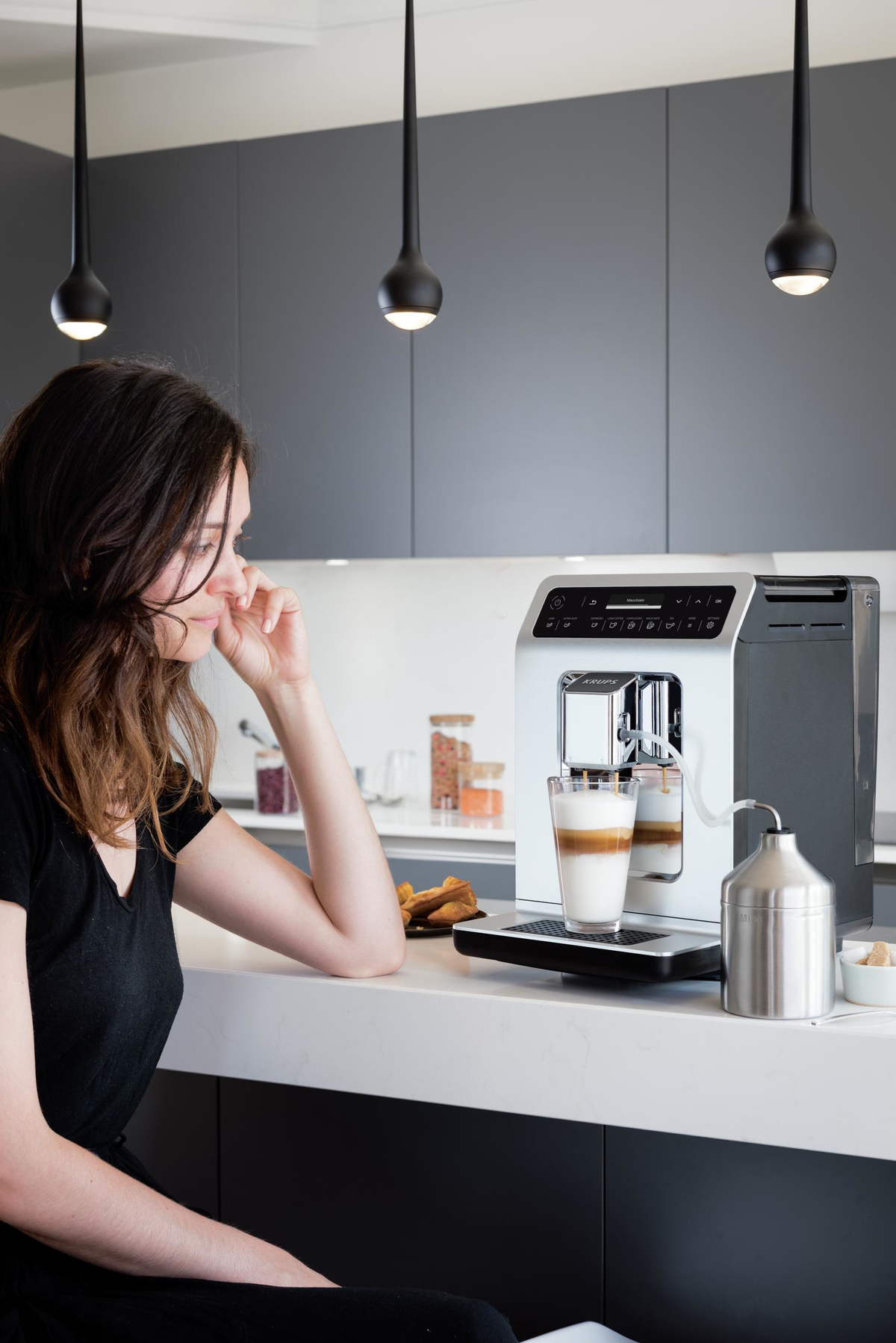 Kobieta siedzi przy ekspresie do kawy. Szklanka kawy z mlekiem na blacie.