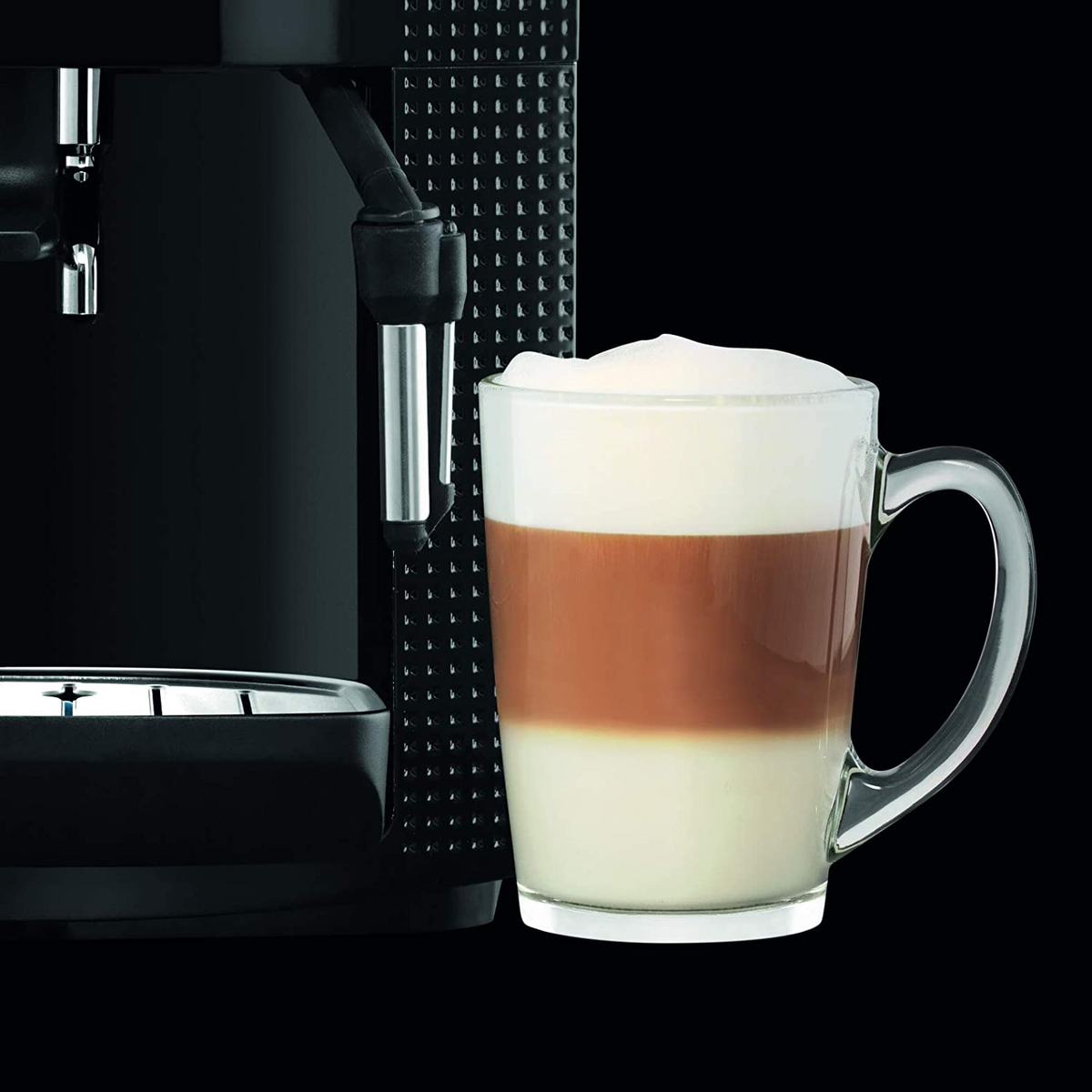 Una tazza di vetro con bevande al caffè a strati accanto a una macchina da caffè nera. Lo sfondo è nero.