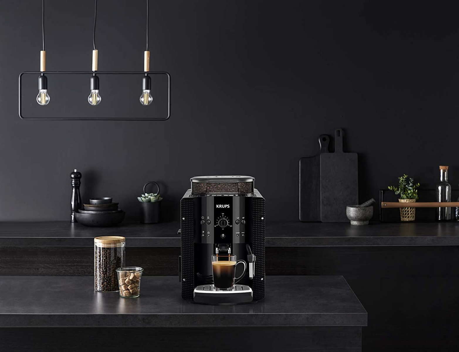 Una macchina per caffè espresso Krups nera è su un piano di lavoro scuro. Ci sono chicchi di caffè, zucchero e un espresso.