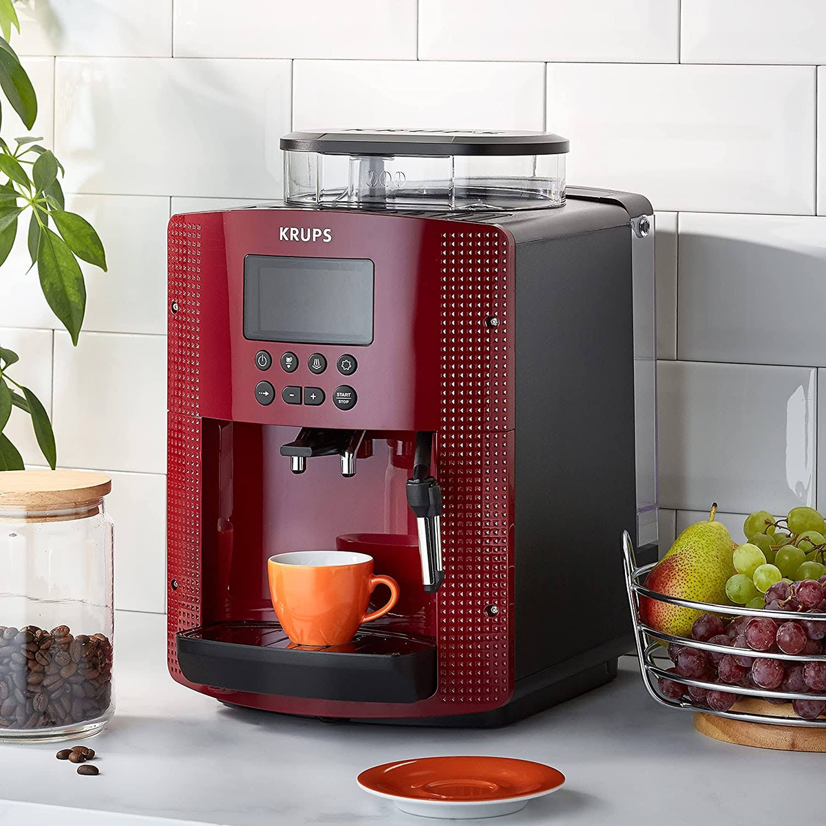 Czerwono-czarny ekspres do kawy Krups. Pomarańczowy kubek, ziarna kawy i kosz owoców na blacie.