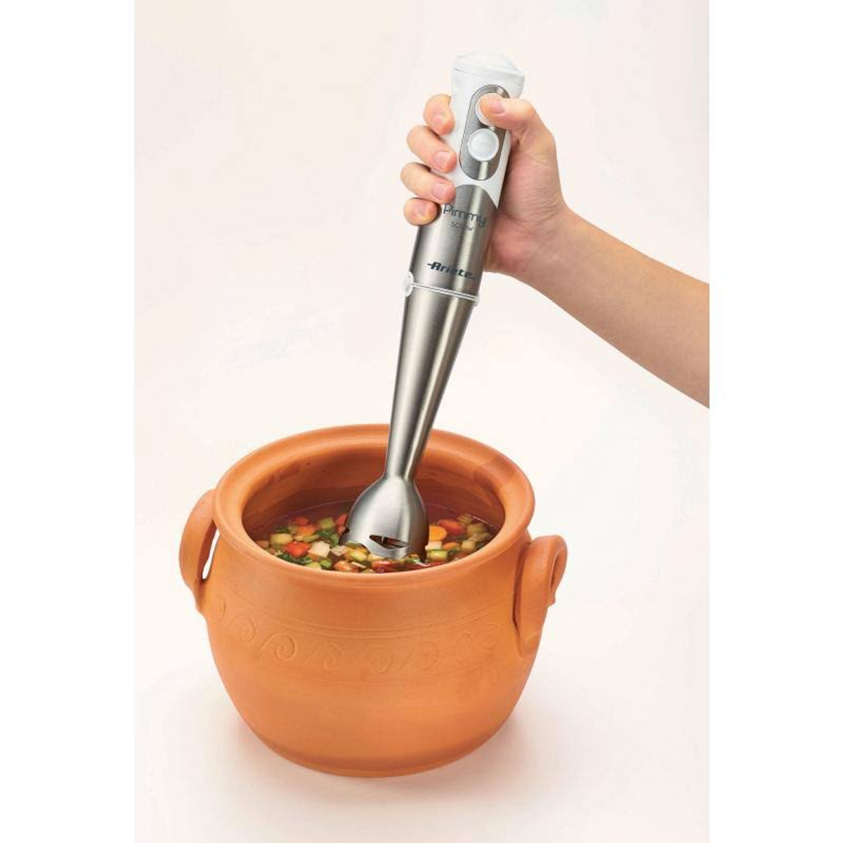 Mano che frulla la zuppa in un vaso di terracotta con un frullatore a immersione argentato. La zuppa contiene verdure.