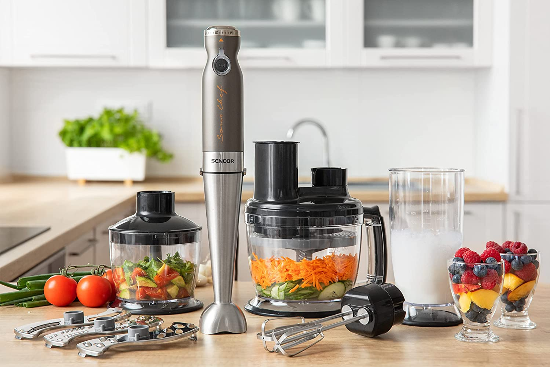 Un set di frullatore a immersione con vari accessori e ingredienti, disposti su un piano di lavoro in legno.