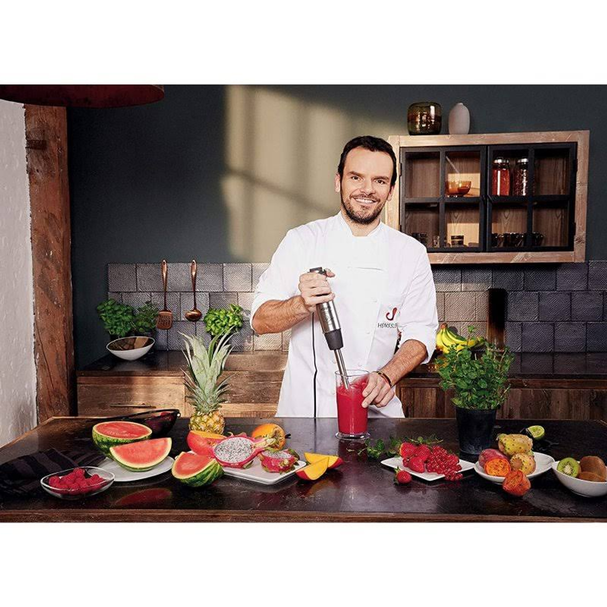 Szef kuchni miksuje czerwony napój blenderem, z owocami i narzędziami na drewnianym stole.