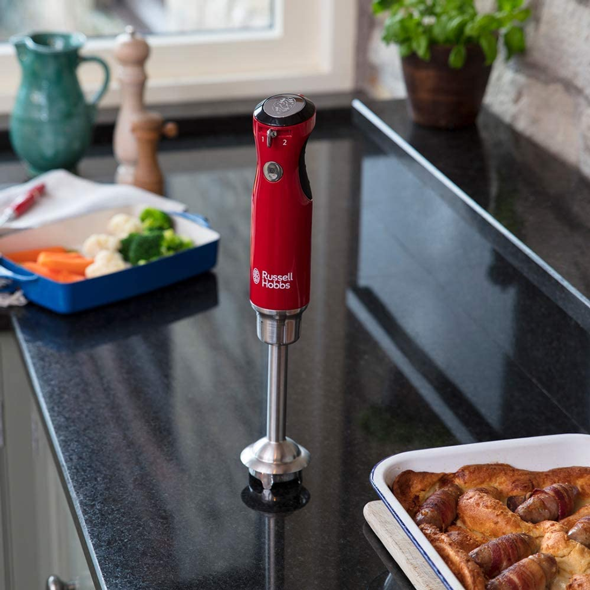 Siyah bir tezgah üzerinde kırmızı bir Russell Hobbs el blenderi. Arka planda yiyecekler var.