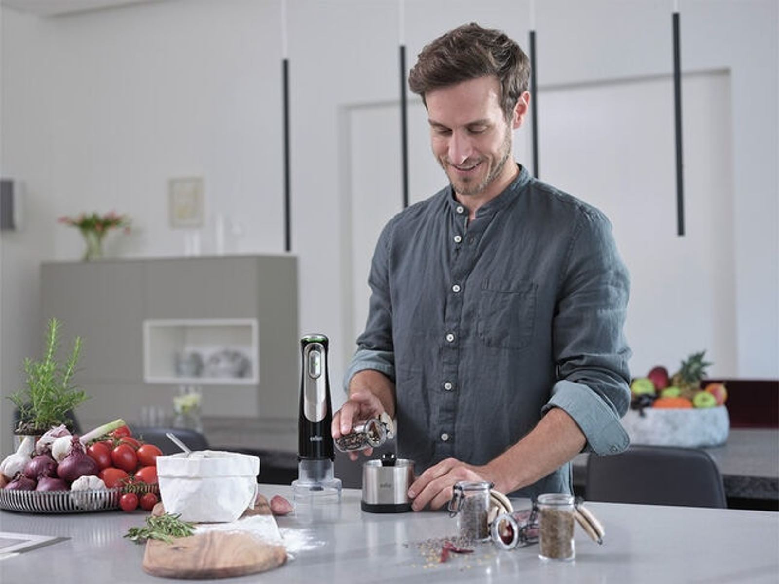 Mężczyzna w szarej koszuli przygotowuje jedzenie. Używa blendera ręcznego i ma różne składniki.